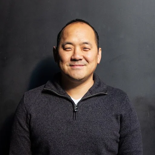 Smiling man with light skin, short hair, and a half-wink, wearing a dark zip-up sweater, standing against a dark background.