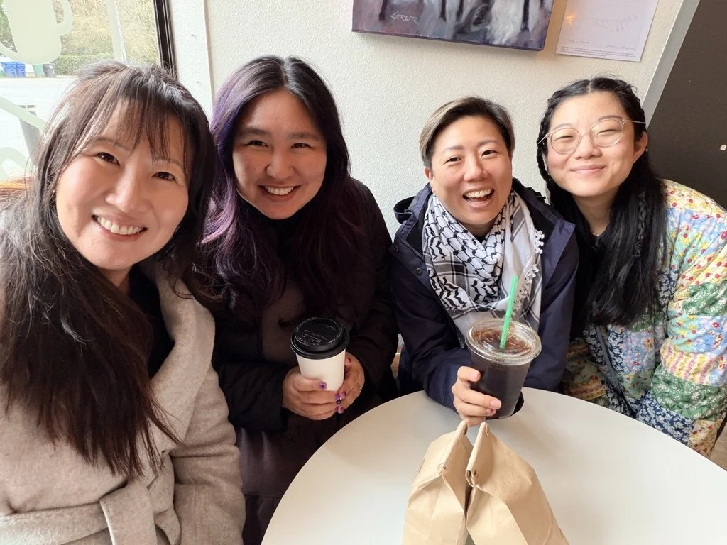 Four women smiling and sitting at a table in a coffee shop, holding drinks, with a window and artwork on the wall in the background.