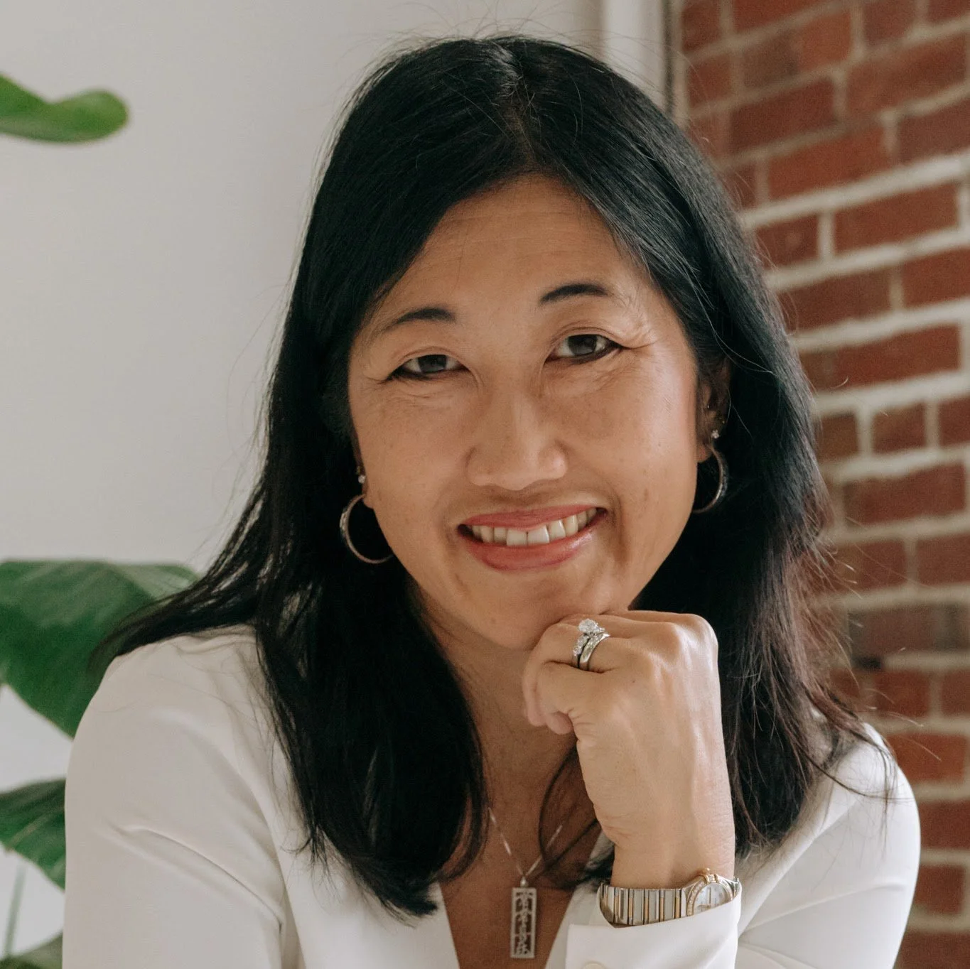 A woman with long dark hair smiling, wearing hoop earrings, a ring, and a watch, sitting in front of a brick wall and a green plant, resting her chin on her hand.