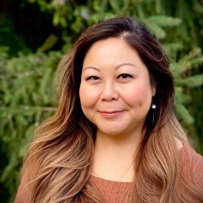 A woman with long, wavy brown hair and earrings, standing outdoors in front of green foliage, smiling softly.