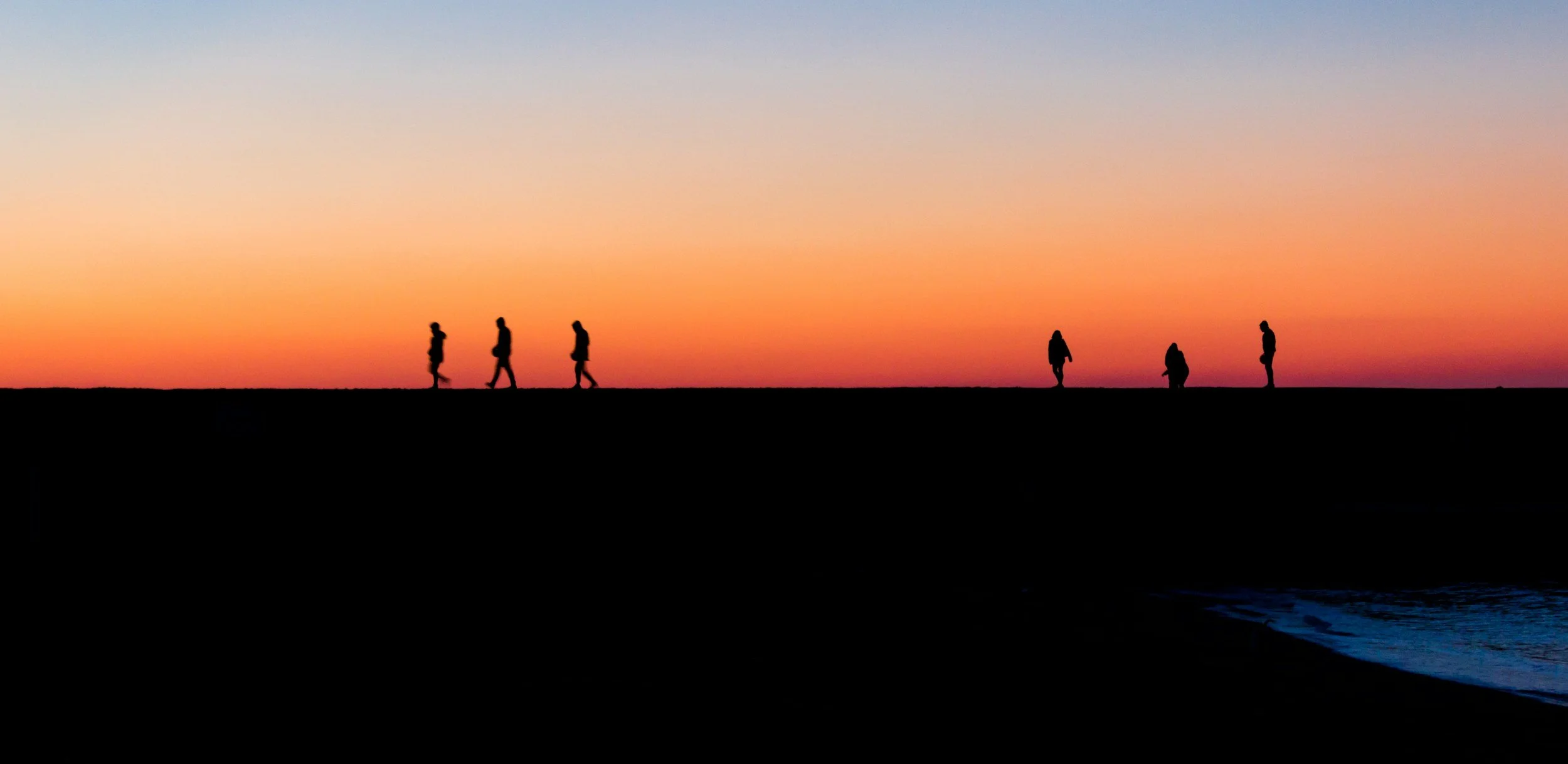Silhouettes of people walking along a beach during sunset with a colorful sky in the background.