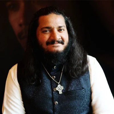A man with long black hair and a beard, smiling, wearing a white shirt and dark vest, with a cross necklace, in a dimly lit setting.