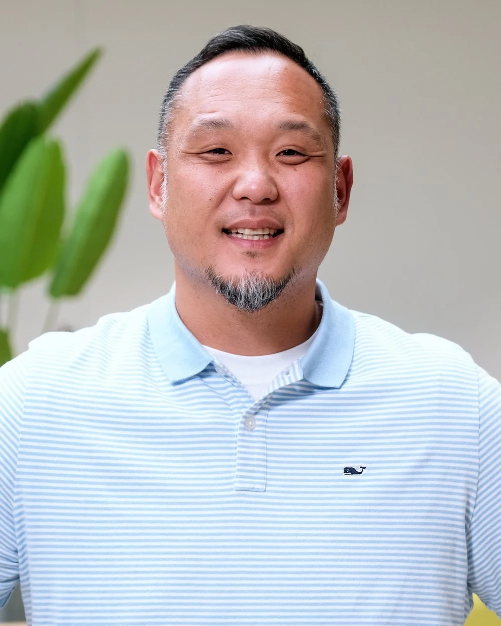 A smiling man with short black hair and a goatee, wearing a light blue striped polo shirt, standing indoors with a blurred plant in the background.