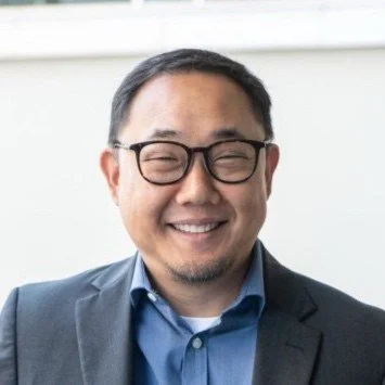 A man in a suit and glasses smiling in an office setting.