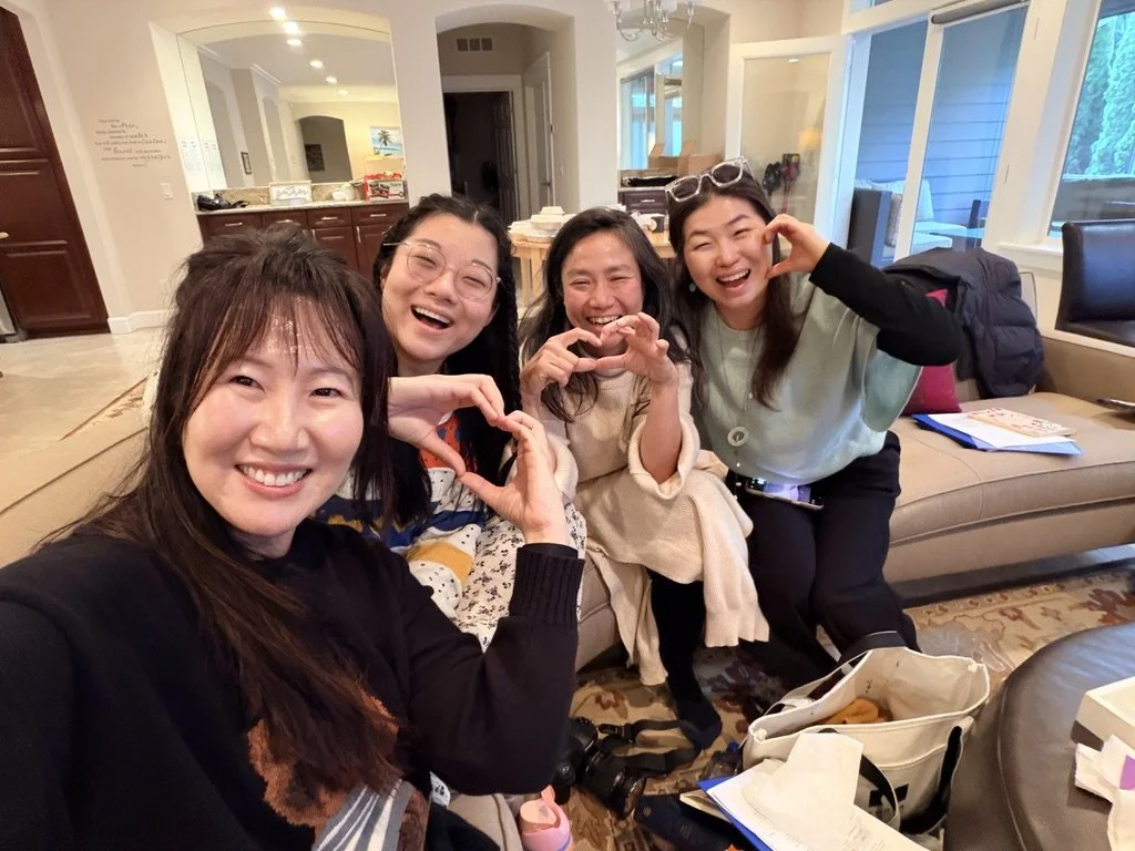 Four women sitting on a beige couch in a living room, smiling and making heart shapes with their hands. There are various items like bags, papers, and a camera in front of them, with a kitchen and windows in the background showing a backyard.