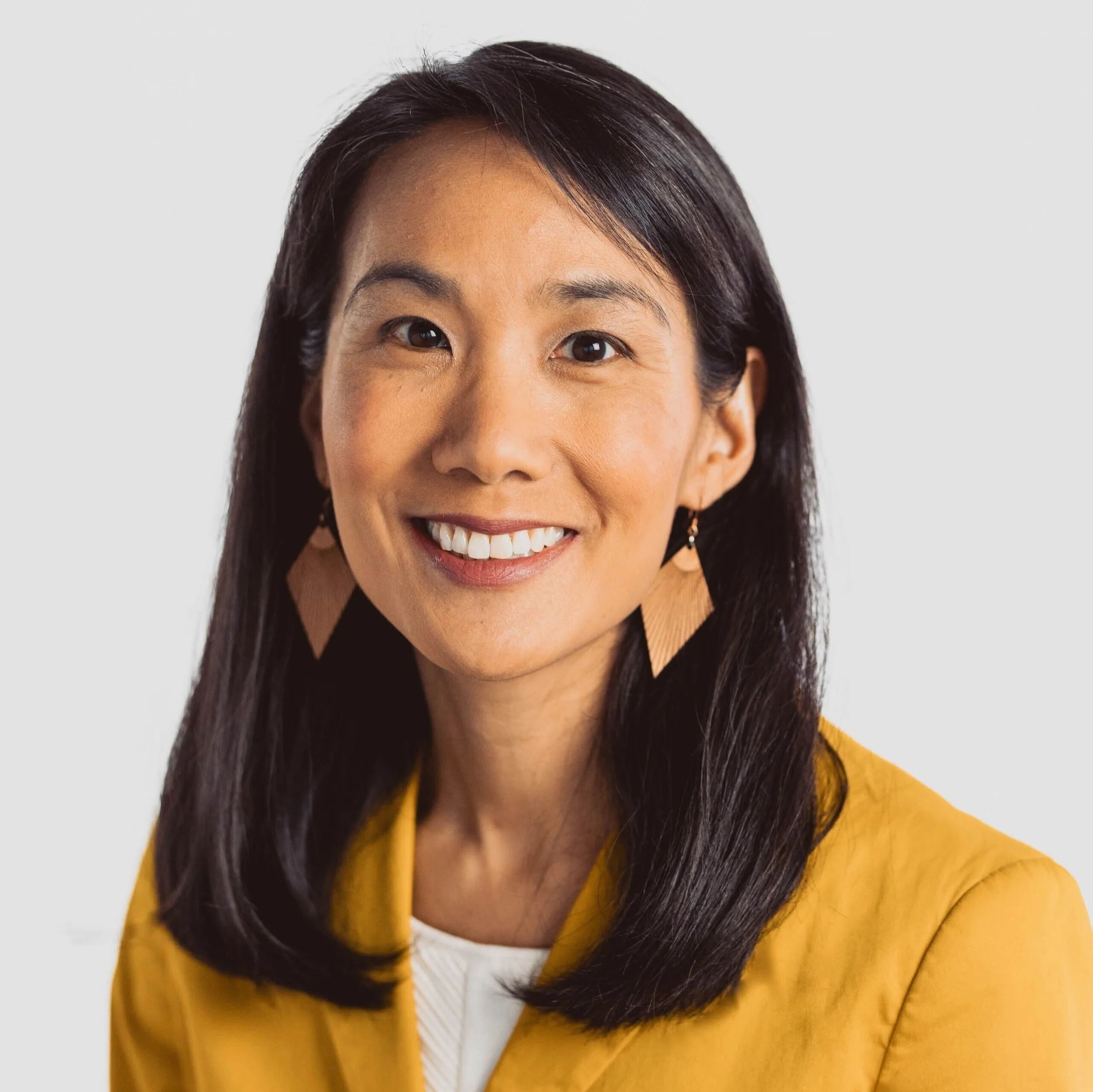 Close-up photo of an Asian woman smiling, with long dark hair, wearing a mustard yellow sweater and earrings, against a neutral background.