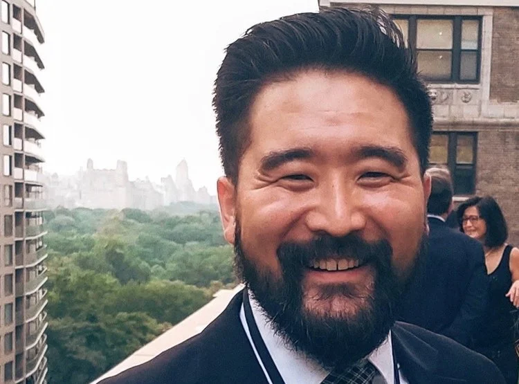 Smiling man with dark hair and beard on balcony with cityscape and trees in background, several people visible in background.
