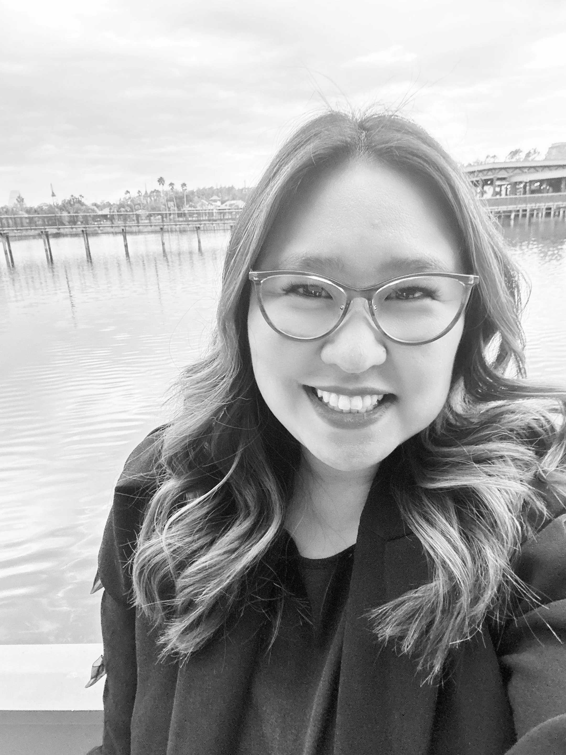A smiling woman with long wavy hair and glasses, taking a selfie outdoors near a body of water with trees and buildings in the background.