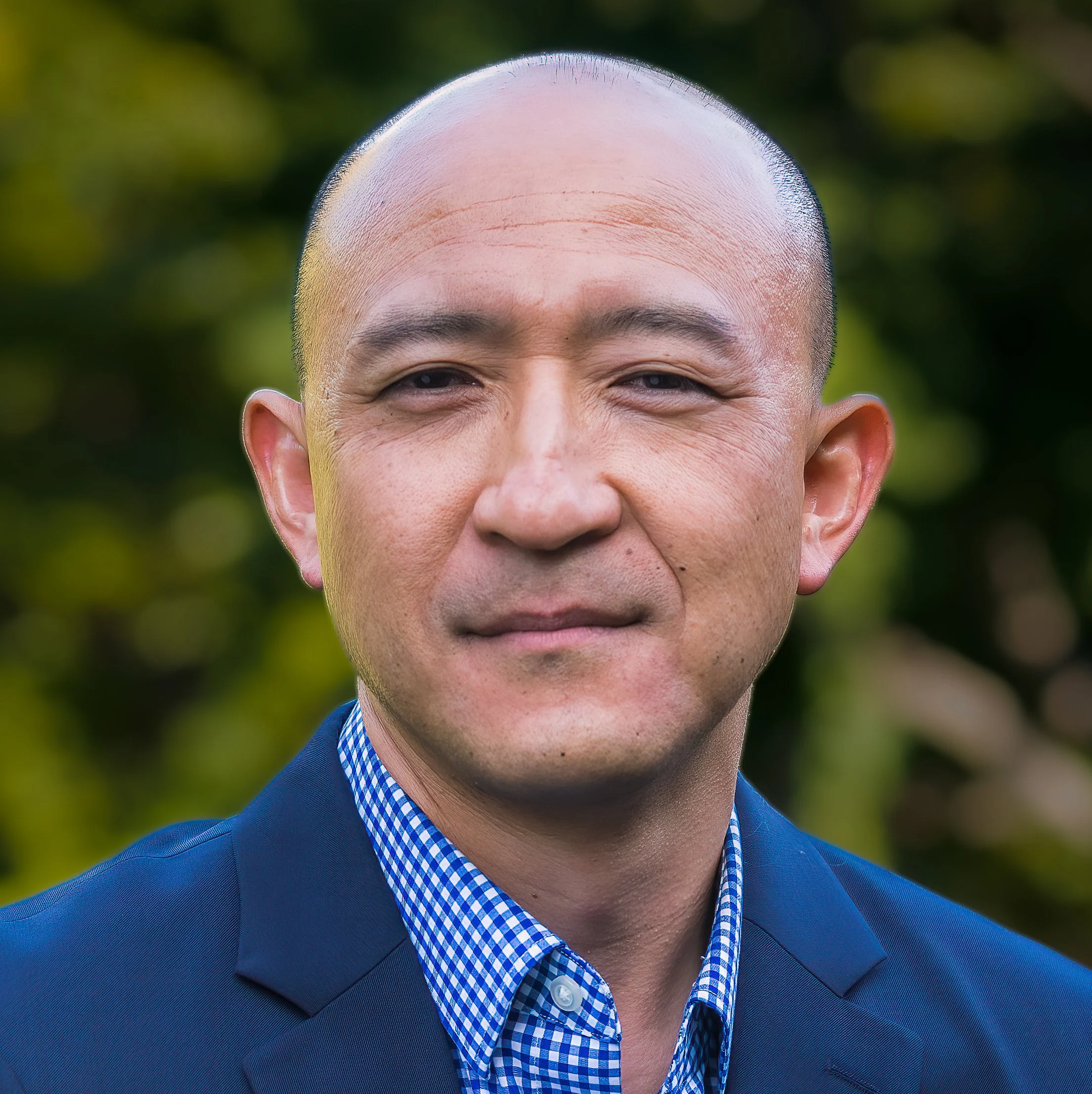 Close-up of a smiling man with a shaved head wearing a blue suit and a blue checkered shirt, outdoors with blurred green foliage in the background.