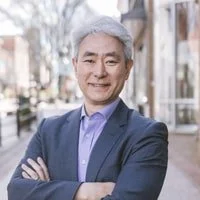 A middle-aged man with gray hair wearing a blue blazer and purple shirt, standing outdoors with an urban background.