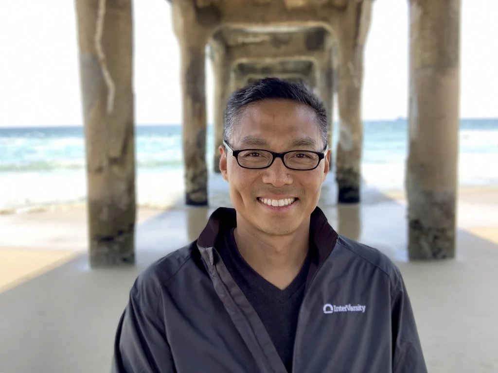 A smiling man with glasses standing under a pier at the beach.