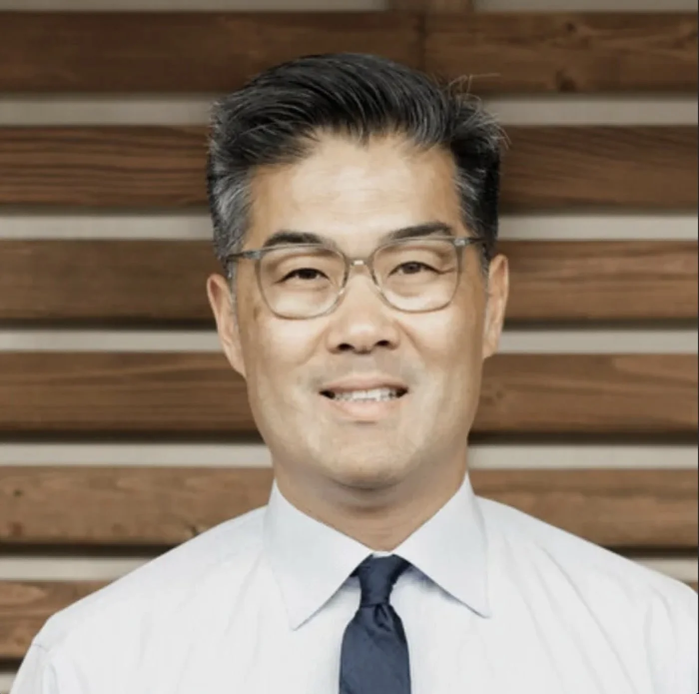 A man with glasses, gray hair, wearing a white shirt and dark tie, smiling in front of a wooden background.