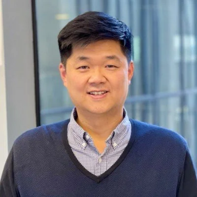 A smiling man with black hair, wearing a gray shirt and a dark blue sweater, standing indoors with large windows in the background.