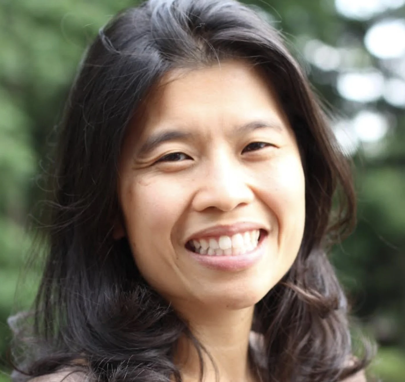 Close-up of a smiling woman with long dark wavy hair outdoors with greenery in the background.