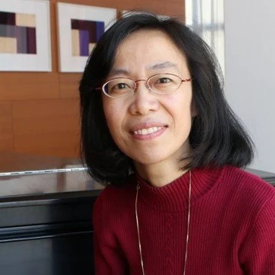A woman with shoulder-length black hair and glasses, wearing a red sweater and a gold chain, smiling at the camera indoors with framed artwork in the background.
