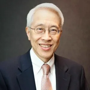 A middle-aged Asian man wearing glasses, a dark suit, a white shirt, and a pink tie, smiling against a plain dark background.