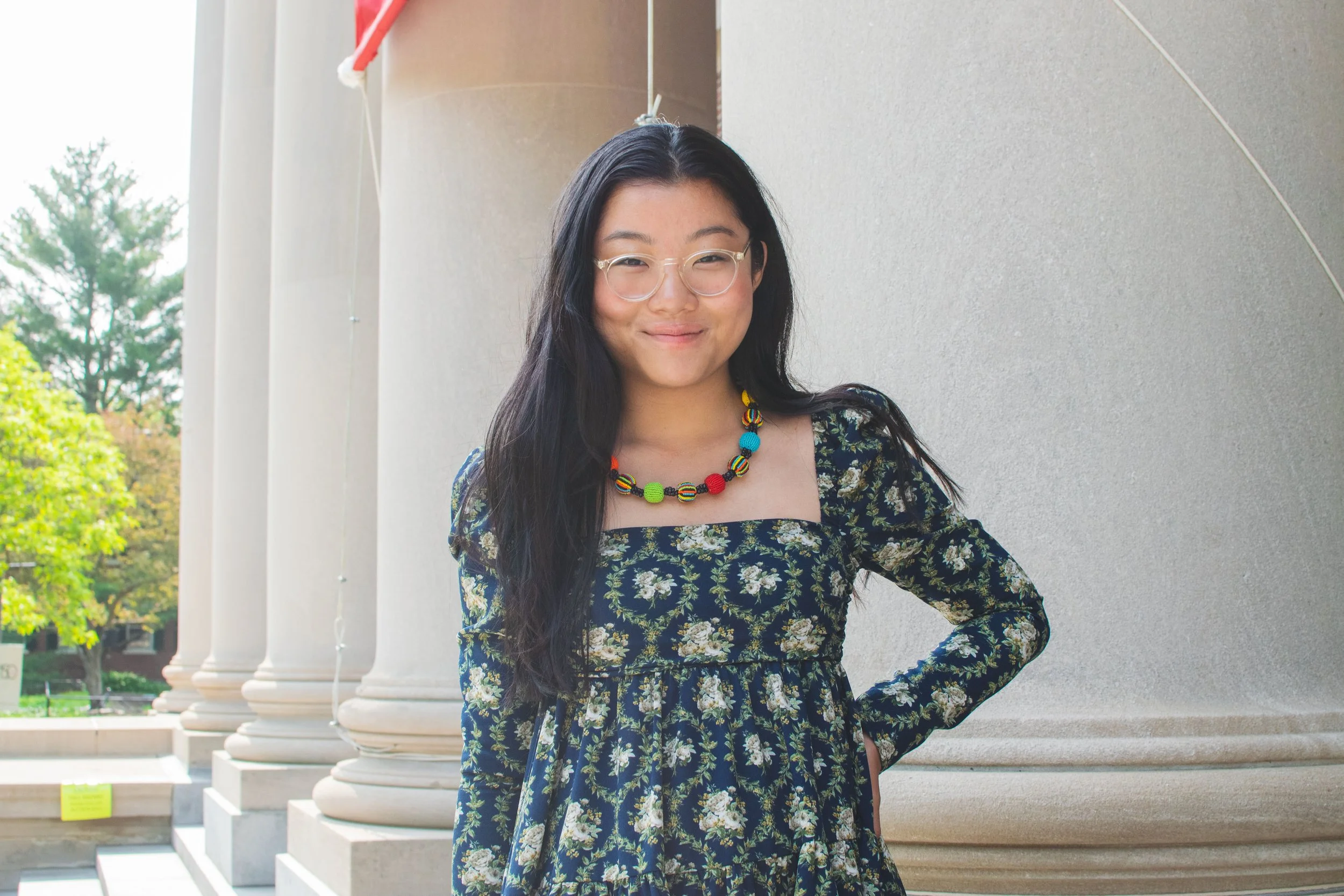 A young woman with long black hair and glasses smiling outdoors near white columns, wearing a floral dress and a colorful beaded necklace.