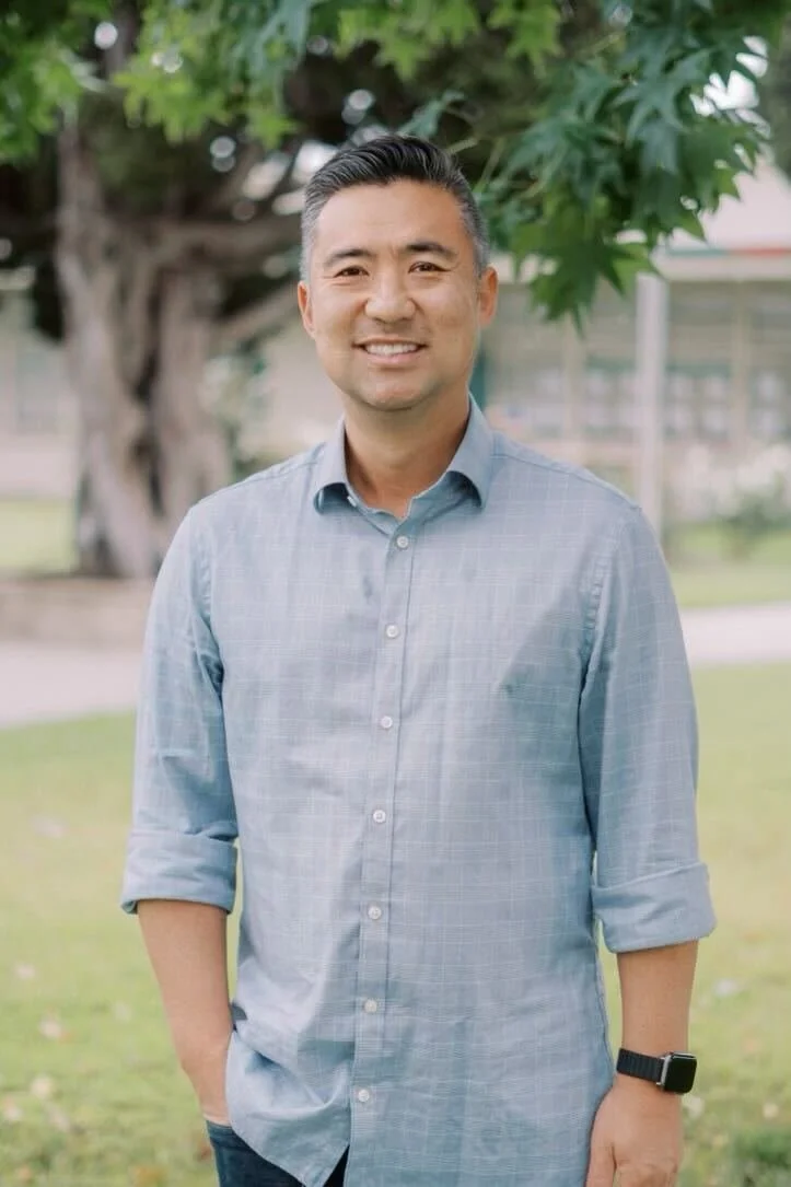 A man stands outdoors in front of a tree and greenery, smiling at the camera, wearing a light blue button-up shirt with rolled-up sleeves and a smartwatch.