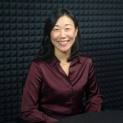 A woman with shoulder-length black hair and a maroon satin shirt, smiling and sitting in front of a black soundproofing wall.