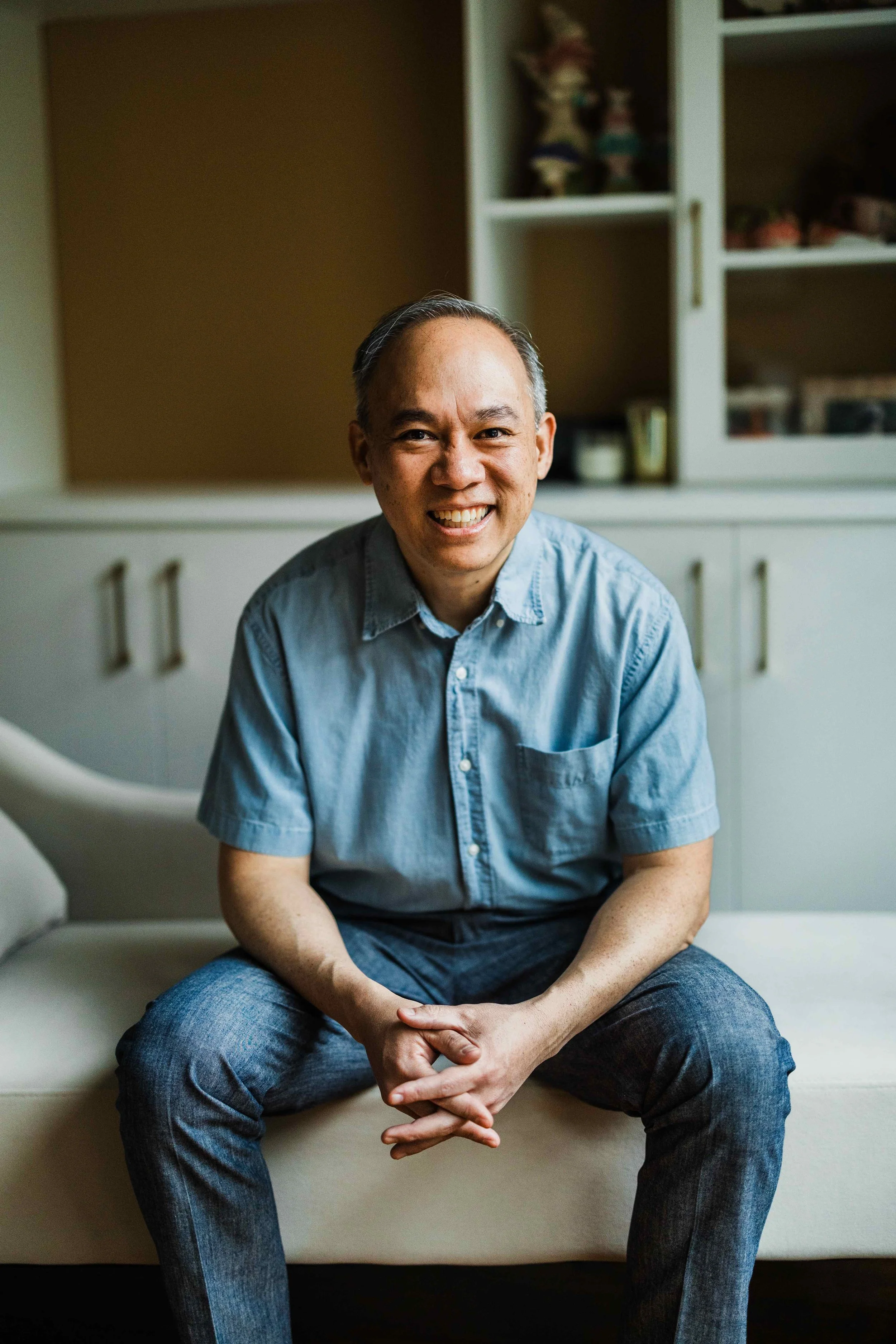 A smiling man with short gray hair wearing a light blue button-up shirt and jeans, sitting on a beige couch in a room with a white bookshelf in the background.