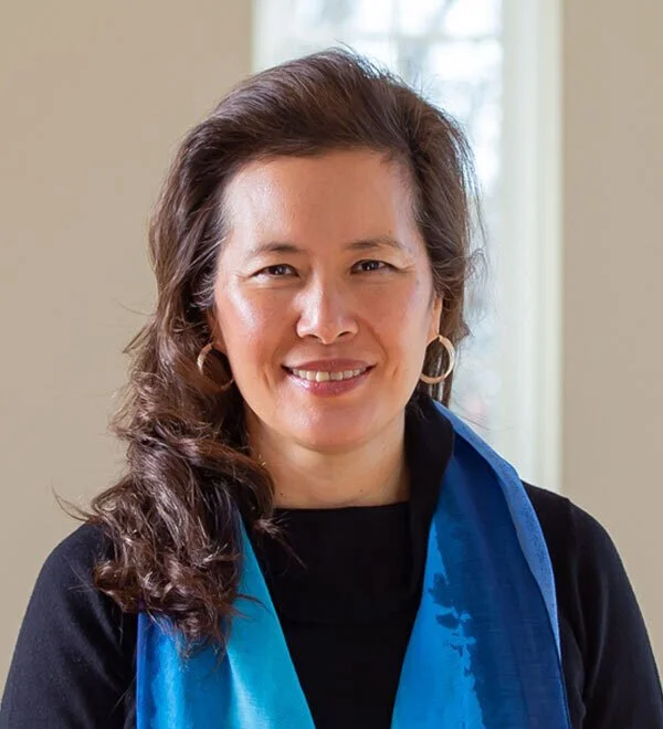 Smiling woman with wavy dark hair, wearing hoop earrings and a blue scarf, standing indoors near a window.