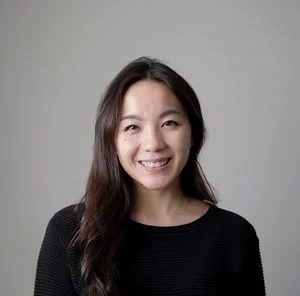 A woman with long dark hair smiling at the camera, wearing a black top against a plain gray background.