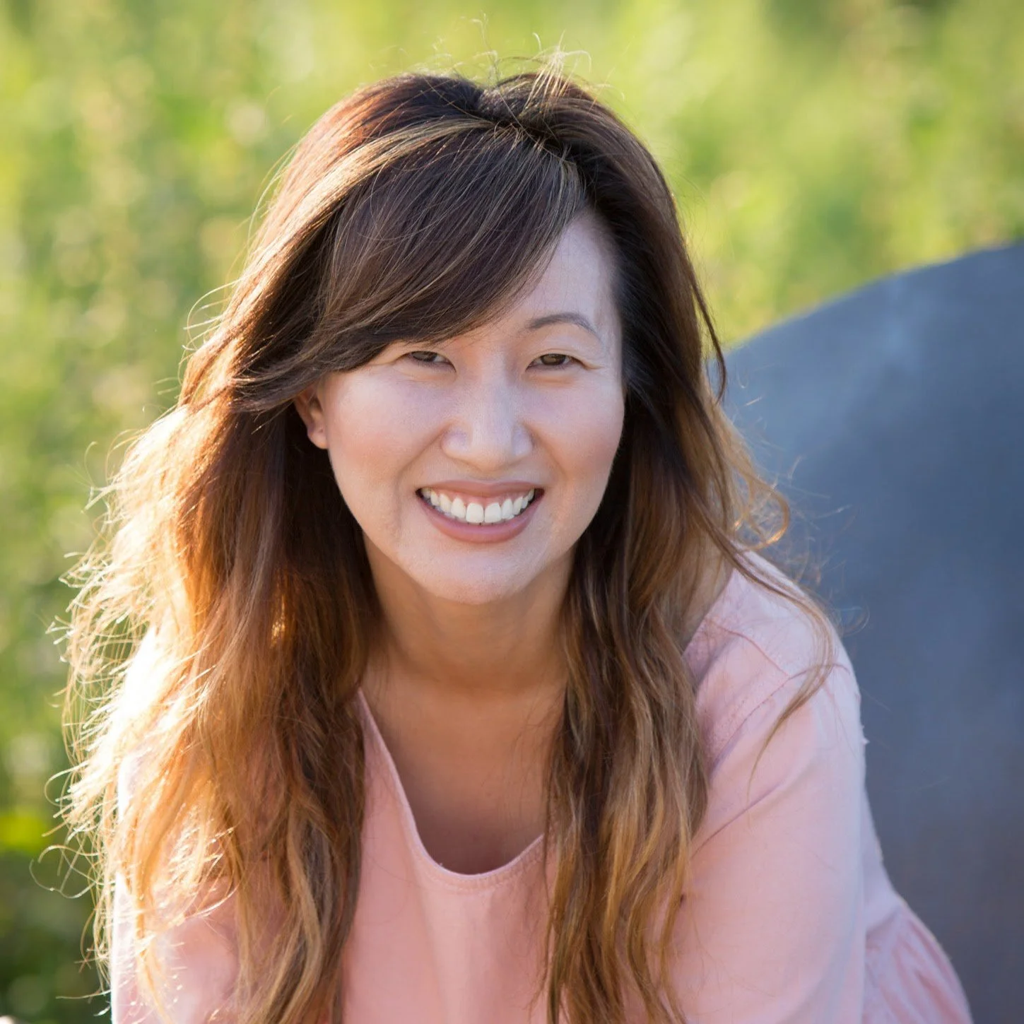 A woman with long wavy hair smiling outdoors in natural light.