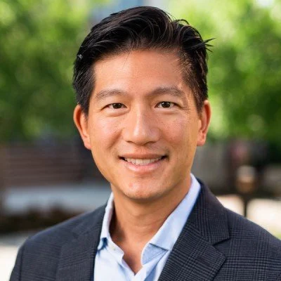 A man in a suit smiling outdoors with trees in the background.