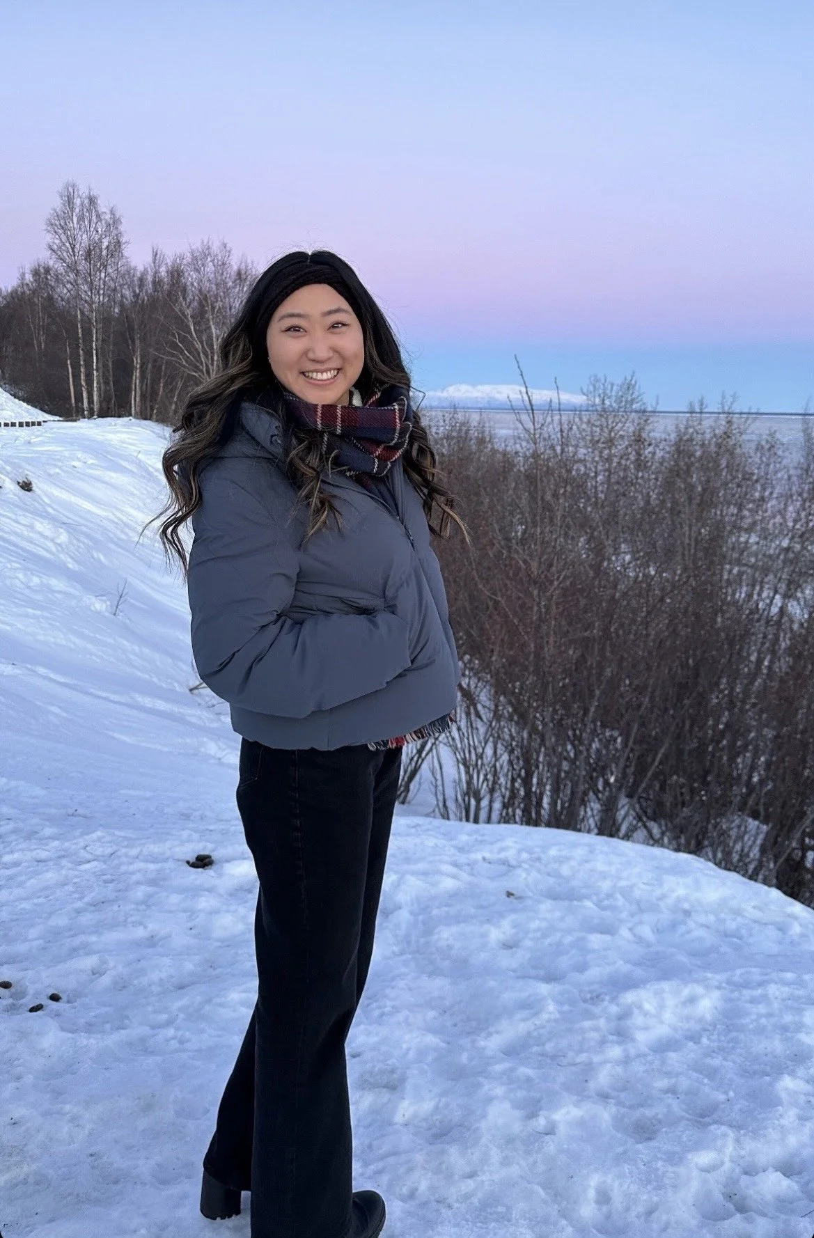 A woman standing in snow-covered landscape during sunset, wearing a gray jacket and a plaid scarf, smiling and looking at the camera.