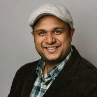 Portrait of a smiling man wearing a gray hat, black blazer, and plaid shirt against a neutral gray background.