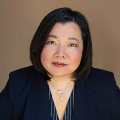 A woman with shoulder-length black hair wearing a dark blazer and a necklace, posing for a portrait against a plain background.