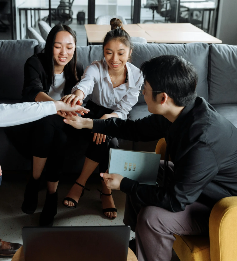 Four young adults participating in a team-building activity in a modern office or coworking space. Two women and one man are on a gray couch; the women are reaching out their hands joined together, and the man is kneeling beside them holding a booklet titled 'I know you girl.' All are smiling and appearing engaged.