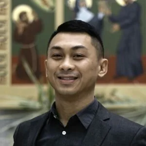 A young man with short black hair smiling, wearing a dark suit, standing in front of a decorative background with depicted figures and objects.