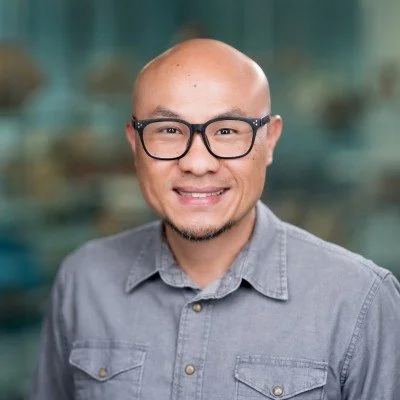A man with glasses, a bald head, and a goatee, smiling and wearing a gray button-up shirt.