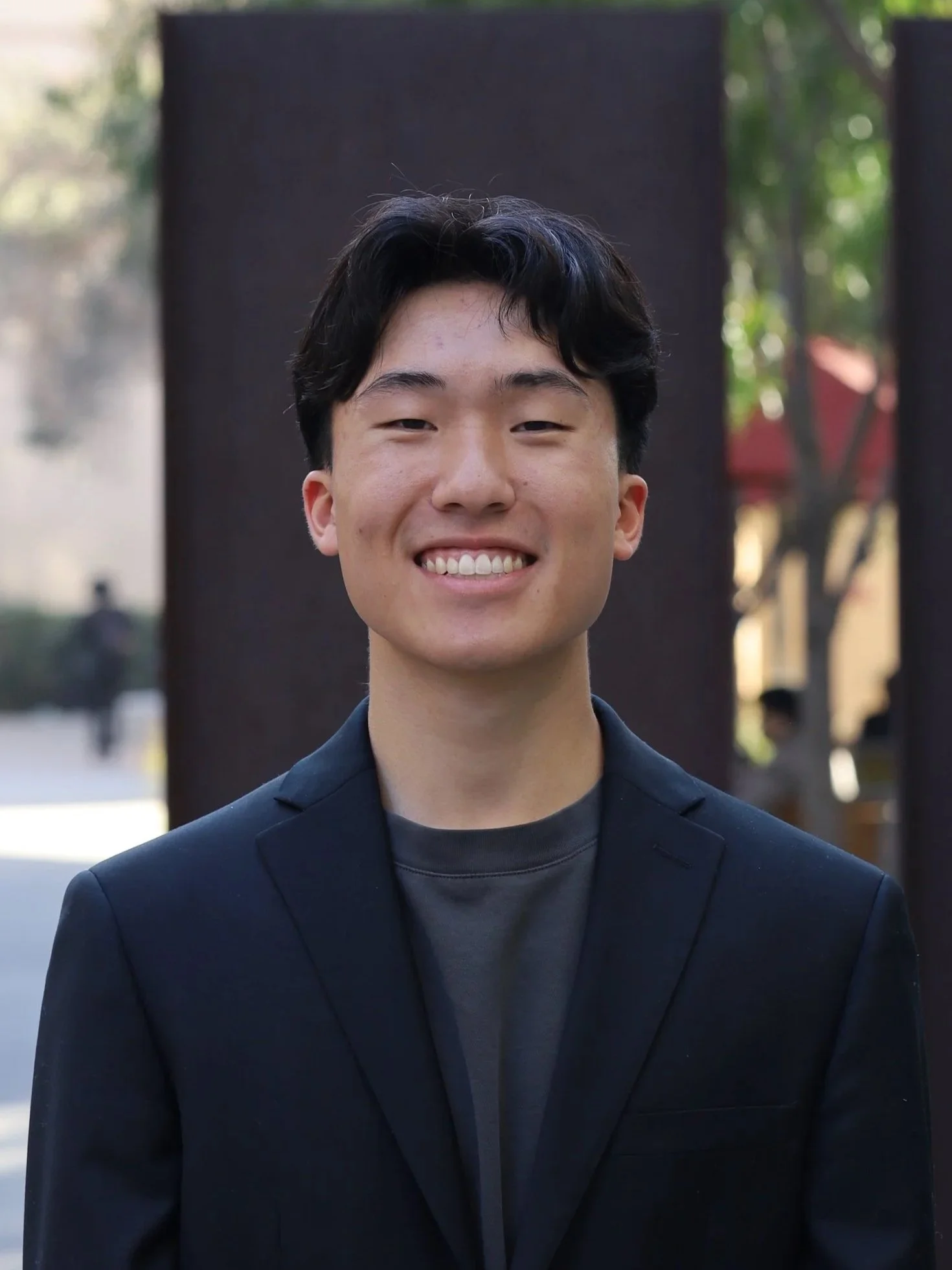 A young man smiling outdoors with trees and buildings in the background, wearing a dark blazer and t-shirt.