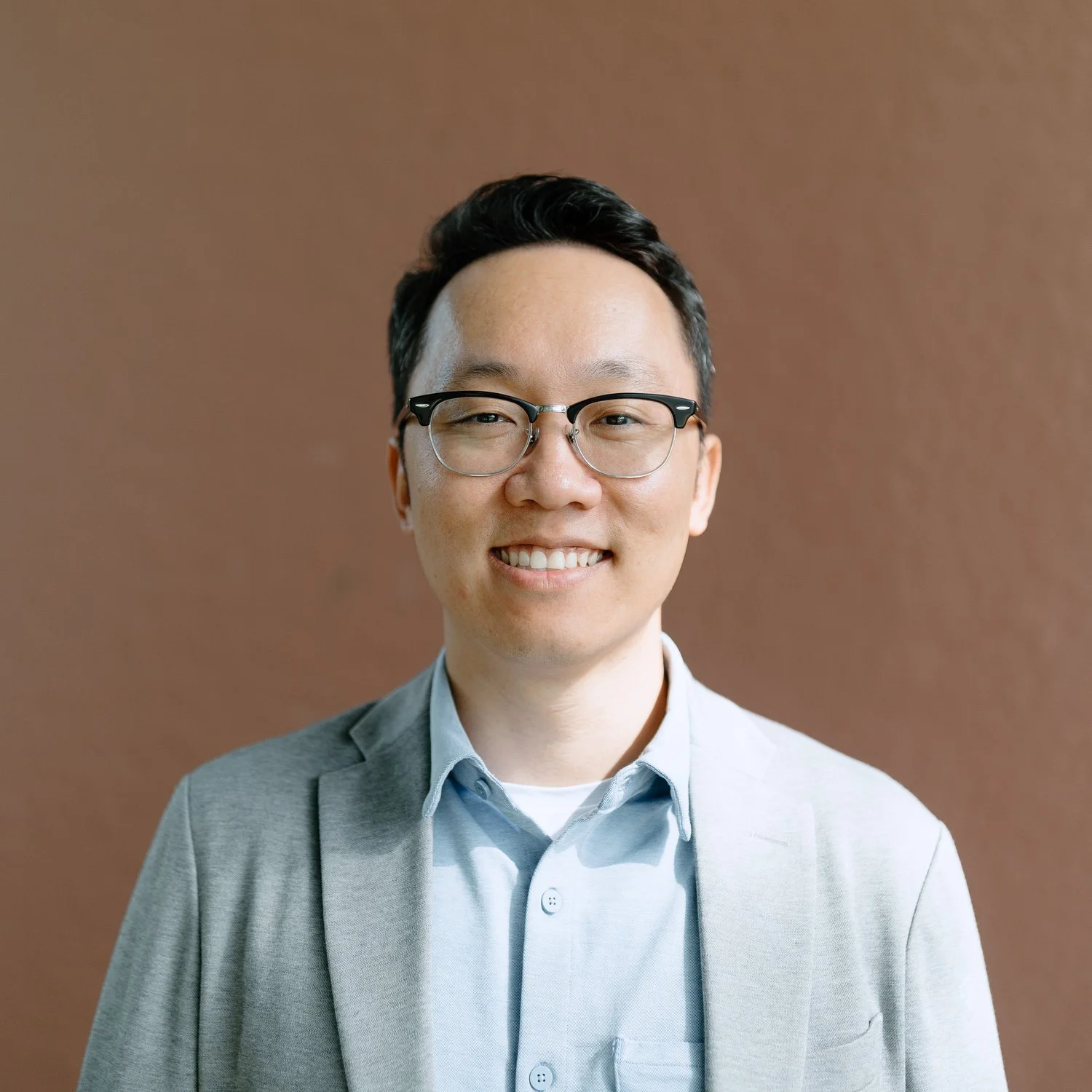 Portrait of a smiling man wearing glasses, a light blue shirt, and a light gray blazer, standing against a brown background.