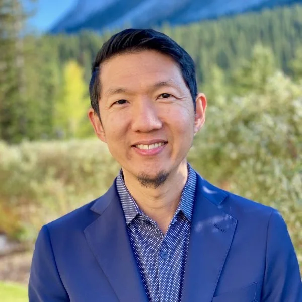 Headshot of a smiling man in a blue suit outdoors with trees and mountains in the background.