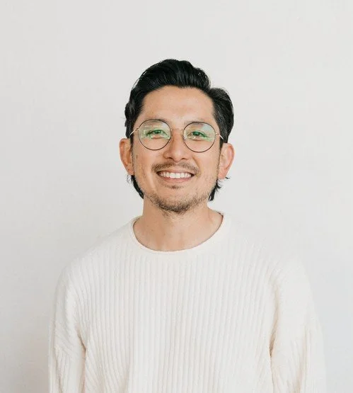 A smiling man with black hair, glasses, and a white sweater against a plain white background.