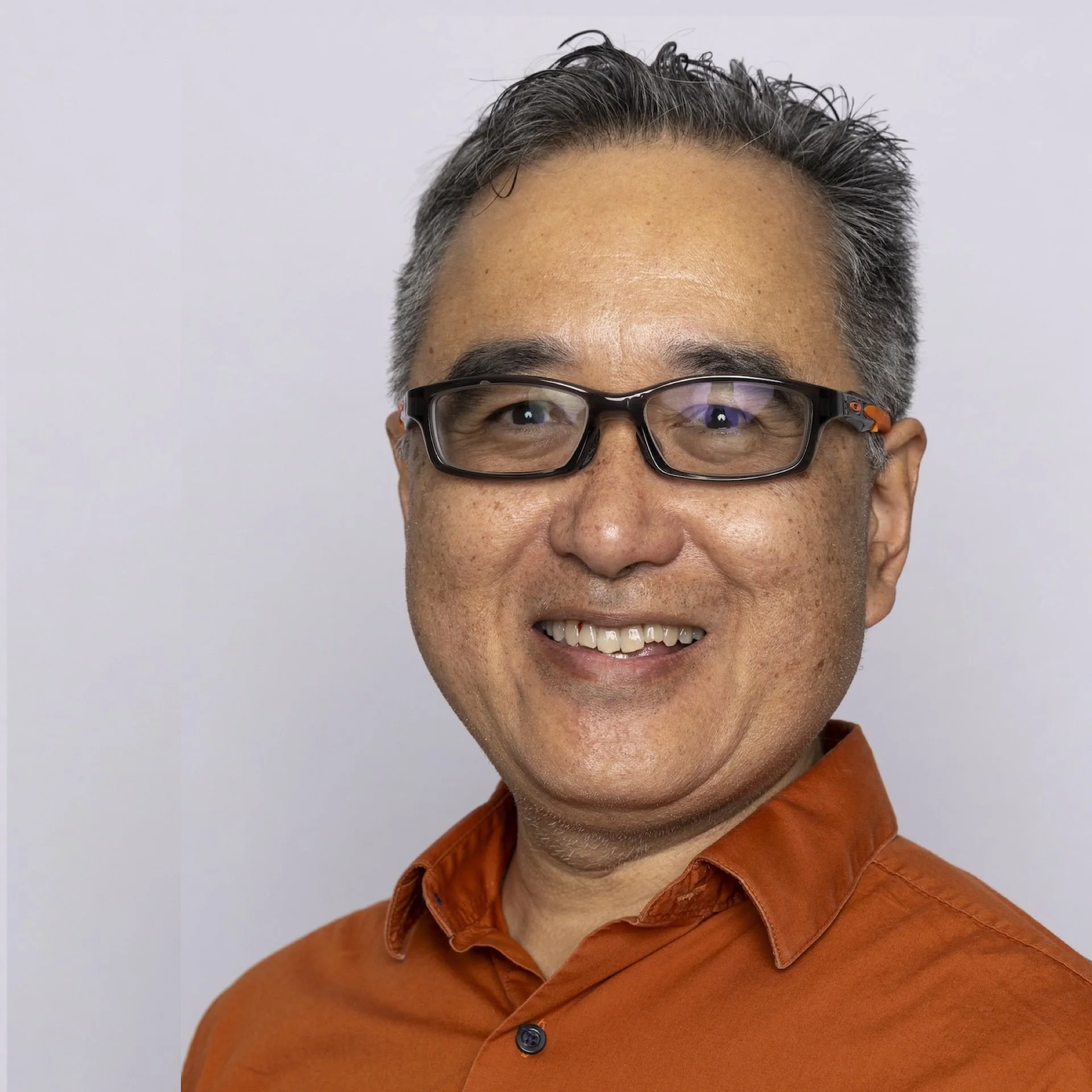 Close-up of a smiling man wearing glasses and an orange shirt against a plain background.