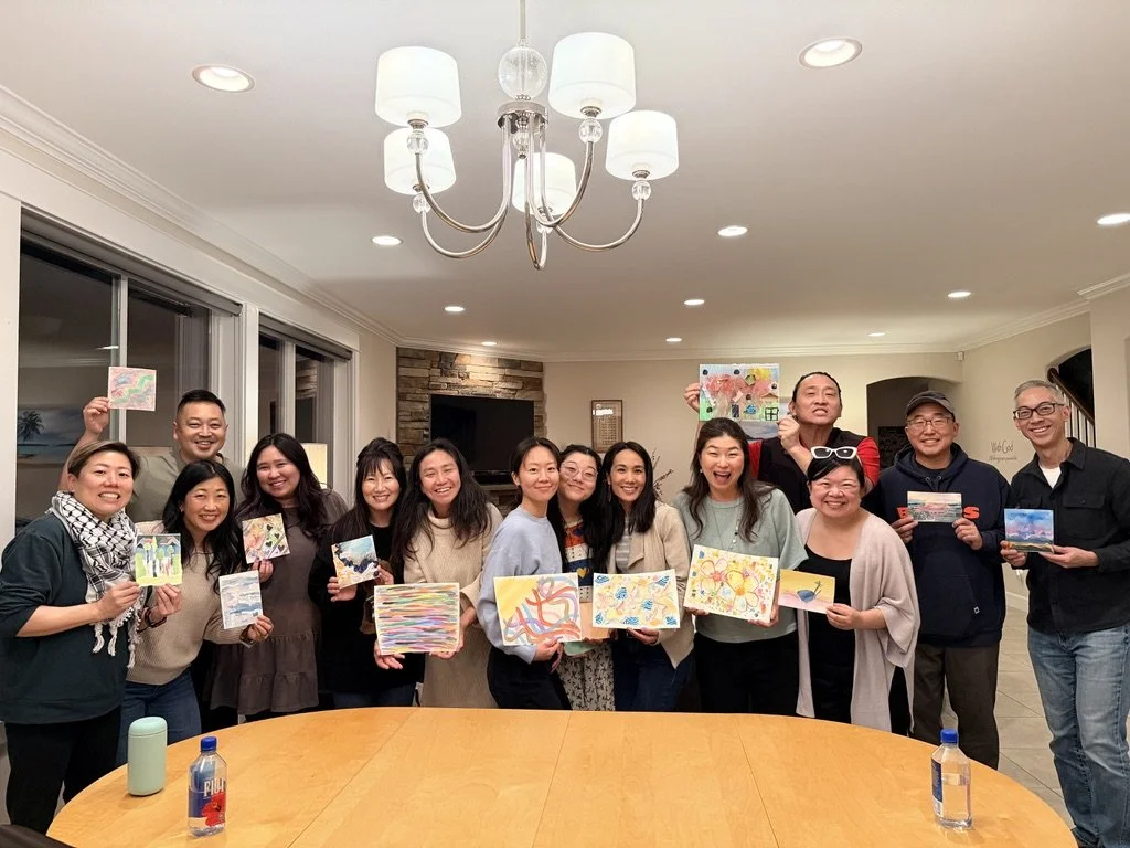 A group of people standing inside a house, each holding a colorful painting they created, smiling at the camera.