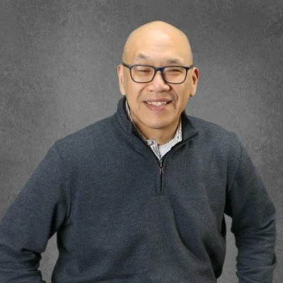 A man with glasses smiling, wearing a dark zip-up sweater over a collared shirt, standing against a dark background.