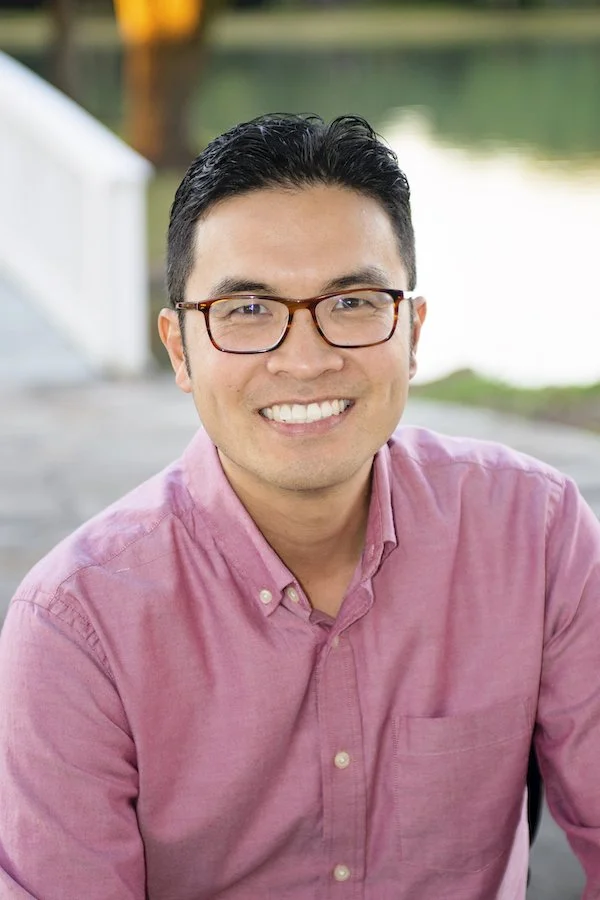 A young man with glasses, wearing a pink button-down shirt, smiling outdoors near water with blurred background.