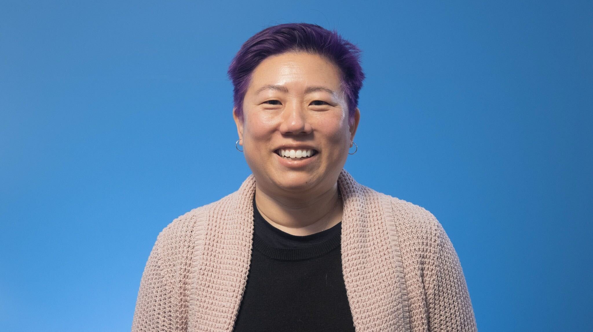 A smiling person with short, styled hair dyed purple, wearing earrings, a black top, and a beige knit cardigan, standing against a blue background.