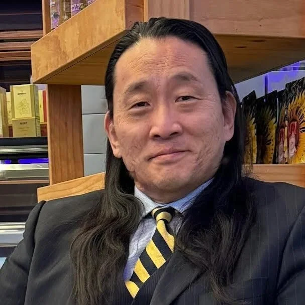 A man with long black hair, dressed in a suit and tie, smiling while seated indoors.