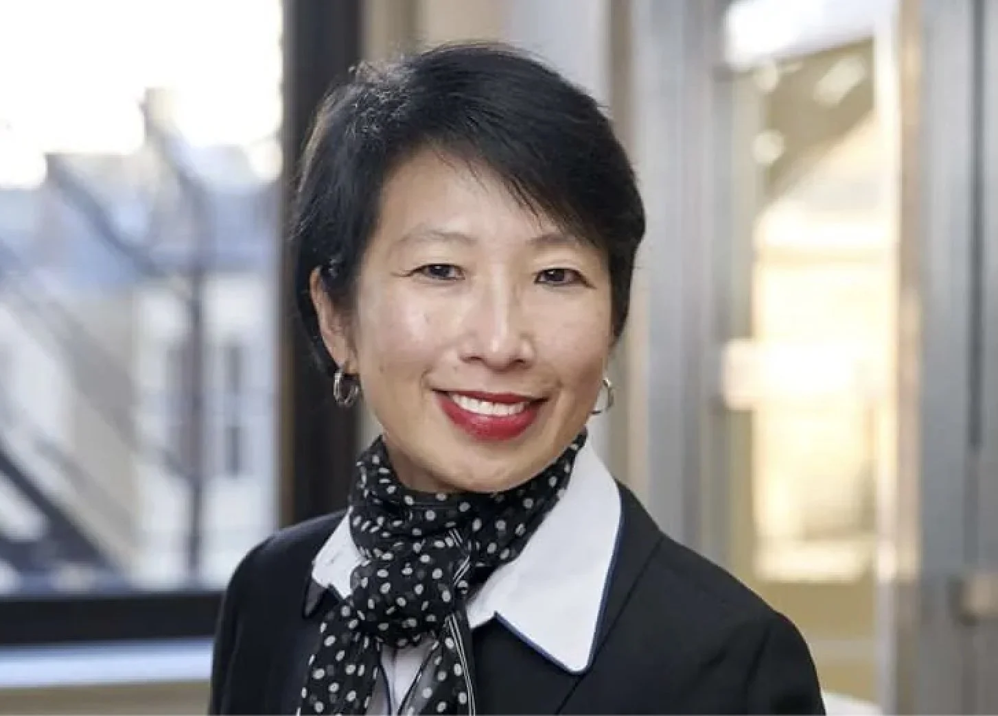 A woman with short black hair and red lipstick smiling, wearing a black blazer, white blouse, and black polka dot scarf, standing in a brightly lit indoor space with windows in the background.