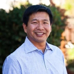 A man smiling outdoors with blurred trees in the background, wearing a light blue collared shirt.