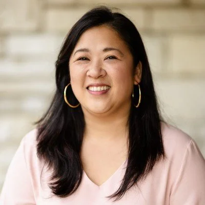 A smiling woman with long black hair, gold hoop earrings, wearing a light pink top, standing outdoors in front of a stone building.