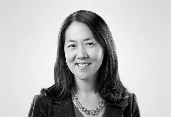 A professional woman with shoulder-length hair, smiling, wearing a dark blazer and a necklace, in a headshot against a light background.