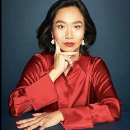 Woman with black hair, red lipstick, and pearl earrings, wearing a red satin blouse, posing with her hand resting on her chin against a dark background.