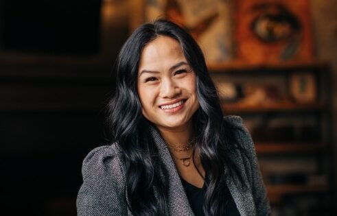 Young woman with long black hair and a bright smile, wearing a gray blazer, in a cozy indoor setting.